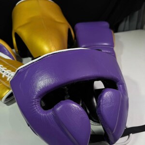 May include: A close-up of boxing headgear and gloves. The headgear is gold and purple, with white trim. The gloves are purple with white trim. The items are made of leather and are designed for protection during boxing or sparring.