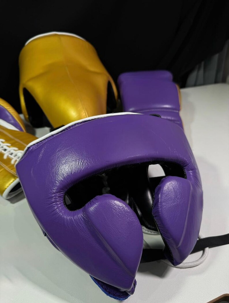 May include: A close-up of boxing headgear and gloves. The headgear is gold and purple, with white trim. The gloves are purple with white trim. The items are made of leather and are designed for protection during boxing or sparring.