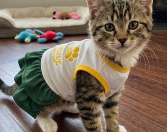 May include: A brown tabby kitten wearing a white mesh tank top with yellow trim and a yellow paw print graphic, paired with a dark green layered skirt. The kitten has white paws and is standing on a dark wood floor with a pet bed in the background.