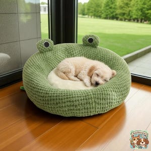 May include: A light green, frog-shaped pet bed with a cream-colored interior. A small, sleeping puppy rests comfortably inside the bed. The bed has two round, textured eyes and is made of a soft, knitted material.