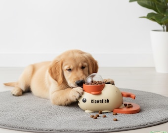 May include: A golden retriever puppy interacts with a beige and orange "Clownfish" dog toy. The toy has a clear, round top and dispenses kibble. The puppy is lying on a gray mat, with scattered kibble around the toy.