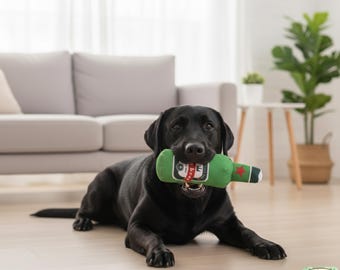May include: A black Labrador Retriever dog is lying down, holding a green plush toy shaped like a beer bottle in its mouth. The toy has a white label with red and black text and a red star. The dog is in a living room setting.