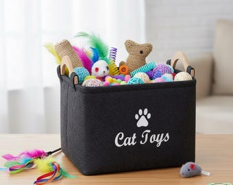 May include: A black fabric cat toy storage bin with wooden handles, filled with colorful cat toys. The bin has a white paw print and the words "Cat Toys". A feather toy and a small gray mouse toy are on the table.