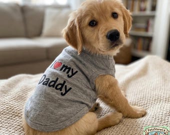 May include: A golden retriever puppy wearing a heather gray sleeveless shirt. The shirt features the text I heart my Daddy printed in black, with a small red heart symbol replacing the word heart. The puppy has light golden fur and dark eyes.