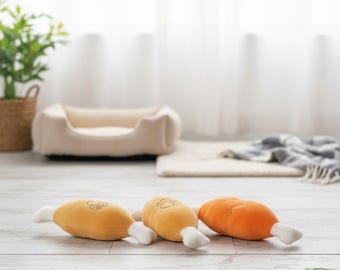 May include: Three plush dog toys shaped like chicken drumsticks in shades of yellow and orange with white bone ends. The toys are on a light wood floor, with a dog bed and plant in the background.