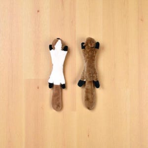 May include: Two plush toy weasels. One weasel is white with a brown head and tail, and black paws. The other weasel is brown with black paws. Both toys are lying on a light brown wooden surface.
