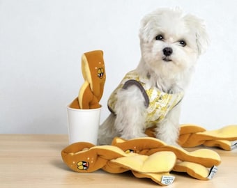 May include: A small white dog wearing a yellow and gray sweater sits near several plush dog toys shaped like pretzels. The toys are brown and yellow with embroidered details. One toy is in a white cup.