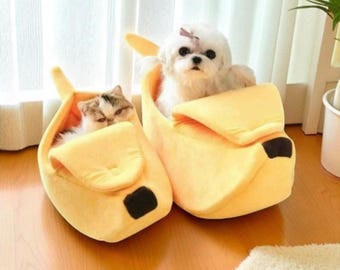 May include: Two small pets, a dog and a cat, are nestled in yellow banana-shaped pet beds. The beds have black accents and are placed on a wooden floor. The dog has a pink bow.