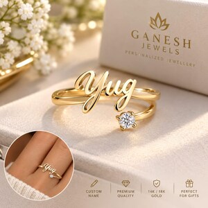 Könnte beinhalten: Ein goldfarbener offener Ring mit dem Namen Yug in Schreibschrift an einem Ende und einem einzelnen runden, klaren Edelstein am anderen Ende. Der Ring wird neben einer Schachtel mit der Aufschrift Ganesh Jewels gezeigt und enthält ein Inset.