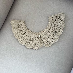 May include: A beige crocheted collar with a scalloped edge and a button fastening. The collar features a delicate, open-weave crochet design. A vintage-style accessory.