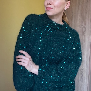 May include: A dark green knit sweater with scattered teal sequins. The sweater has a crew neck and long sleeves. The sequins add a touch of sparkle to the otherwise simple design. The sweater appears to be made of a soft, textured yarn.