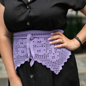 May include: A lilac-colored, openwork crochet belt with a tie closure is worn over a black dress. The belt has a scalloped edge and is tied at the front. The dress has a button-down front and short sleeves.