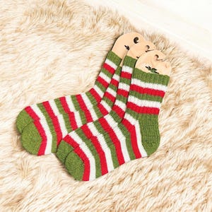 Puede incluir: Un par de calcetines a rayas verdes, rojas y blancas. Los calcetines están hechos de material de punto y se exhiben sobre una superficie suave y de color claro. Los calcetines tienen un diseño clásico y festivo.