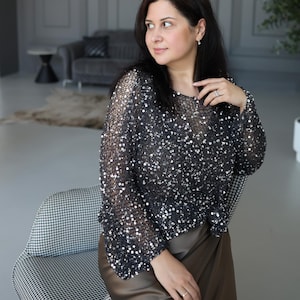 May include: A black sequined top with long sleeves. The top is paired with a brown skirt. The woman is sitting on a black and white checkered chair. The background includes a grey sofa and a side table.