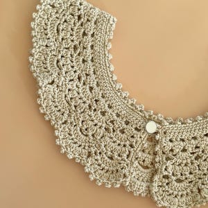 May include: A beige crocheted collar with a scalloped edge and a button fastening. The collar's crochet pattern is intricate and open. This handmade accessory is a neutral colour and would suit many outfits.