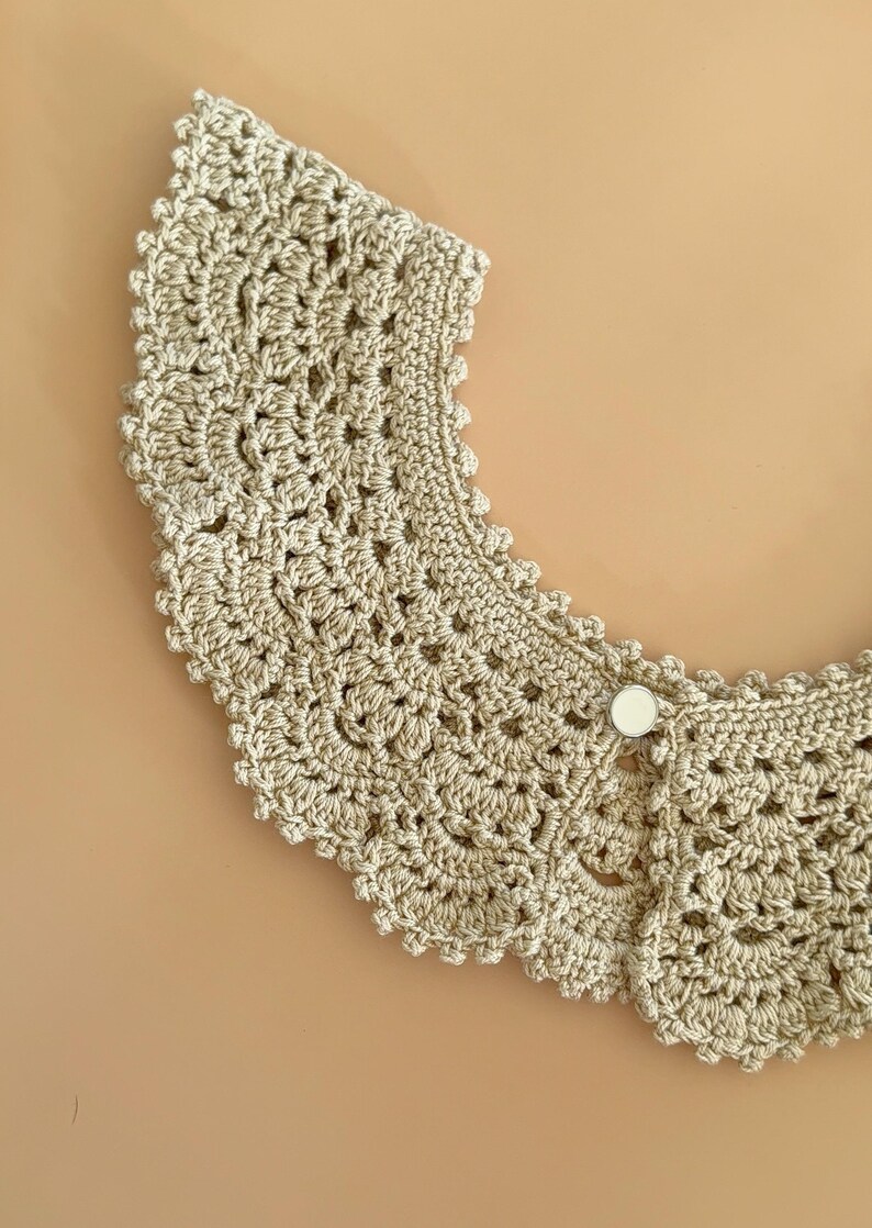 May include: A beige crocheted collar with a scalloped edge and a button fastening. The collar's crochet pattern is intricate and open. This handmade accessory is a neutral colour and would suit many outfits.