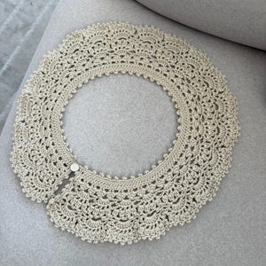 May include: A cream-coloured, crocheted collar with a scalloped edge and a button closure. The collar is circular and appears to be handmade, with intricate openwork detailing throughout. The collar is lying on a grey surface.