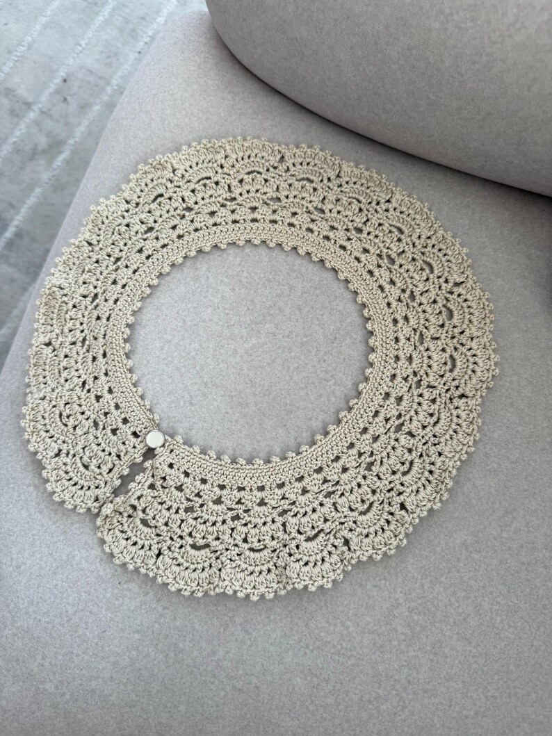 May include: A cream-coloured, crocheted collar with a scalloped edge and a button closure. The collar is circular and appears to be handmade, with intricate openwork detailing throughout. The collar is lying on a grey surface.