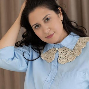 May include: Pale blue button-down shirt featuring a cream-coloured, crocheted collar. The collar has a delicate, scalloped design. The shirt has a single pocket on the left side and is buttoned up. The model is wearing the shirt.
