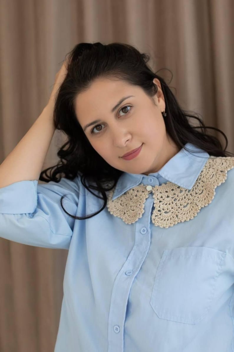 May include: Pale blue button-down shirt featuring a cream-coloured, crocheted collar. The collar has a delicate, scalloped design. The shirt has a single pocket on the left side and is buttoned up. The model is wearing the shirt.