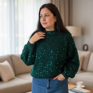 Pode incluir: Um suéter verde escuro com lantejoulas. O suéter tem gola alta e mangas compridas. A pessoa está usando uma saia jeans. O fundo inclui um sofá bege e uma mesa de centro de madeira.