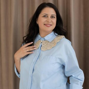 May include: Pale blue button-down shirt featuring a beige crochet collar. The shirt has long sleeves and a single chest pocket. The collar is a delicate, openwork design. The woman is wearing black trousers.