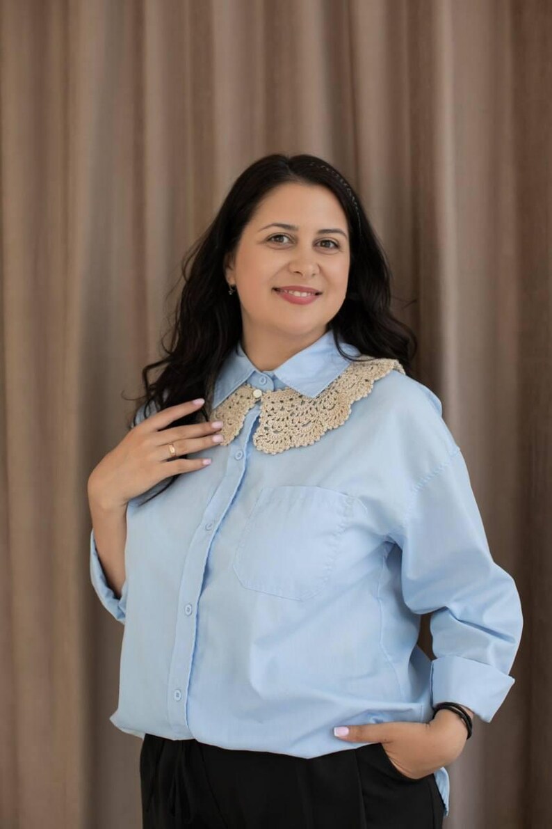 May include: Pale blue button-down shirt featuring a beige crochet collar. The shirt has long sleeves and a single chest pocket. The collar is a delicate, openwork design. The woman is wearing black trousers.
