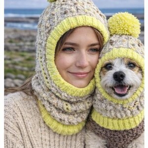 May include: A woman and a small dog are wearing matching knitted hats and neck warmers. The hats have yellow pom-poms and trim. The woman is wearing a beige sweater. The dog is wearing a matching sweater. The background is a blurred outdoor scene.