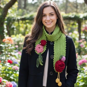 May include: A green crochet scarf with red and pink flower accents. The scarf is adorned with three-dimensional crochet flowers and yellow pom-poms. The scarf is worn over a black coat.