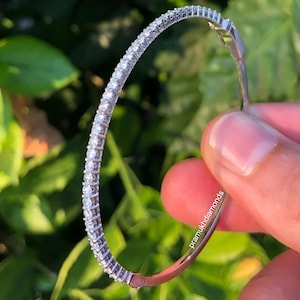 May include: A silver hoop earring with a row of small, clear gemstones. The earring is held in a hand against a backdrop of green foliage. The text "pramukhdiamonds" is visible on the earring.