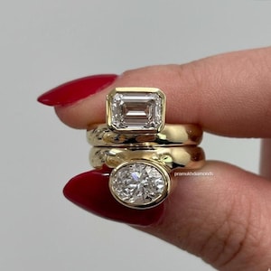 Puede incluir: Tres anillos de oro con piedras preciosas grandes y claras. El anillo superior presenta una piedra de talla esmeralda, el anillo central es una banda lisa y el anillo inferior tiene una piedra de talla ovalada. Los anillos son sostenidos por una persona con esmalte de uñas rojo.