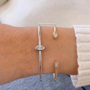 Puede incluir: Tres pulseras de diamantes. Una es un brazalete plateado con un diamante en forma de marquesa en el centro. Las otras dos son pulseras abiertas doradas, cada una con un diamante en forma de marquesa. Las pulseras se llevan en una muñeca.