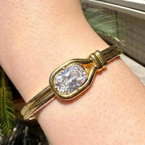 May include: A gold-toned bracelet featuring a large, oval-cut diamond. The diamond is set in a bezel setting, and the bracelet has a unique design with a knot detail. The brand name "pramukhdiamonds" is engraved on the bracelet.
