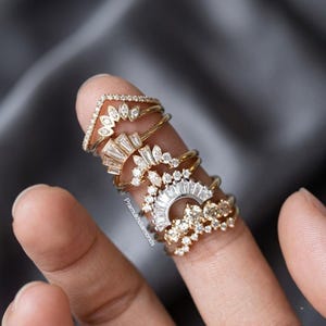Puede incluir: Una pila de seis anillos de oro y plata con varios engastes de diamantes, incluyendo piedras de talla marquesa, baguette y redonda. Los anillos muestran intrincados diseños geométricos con piedras claras y brillantes y el texto Pramukhdiamonds.