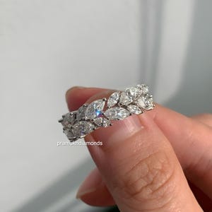 May include: A close-up of a diamond ring featuring marquise-cut stones. The ring has a double-row design, with the diamonds set closely together. The ring is silver in color and reflects light, creating a sparkling effect. The text "pramukhdiamonds" is visible.