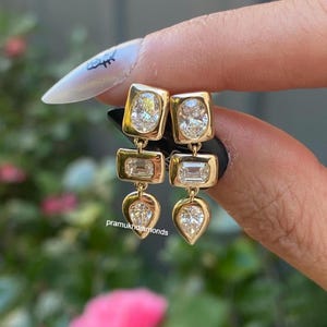 May include: A pair of gold drop earrings featuring three bezel-set diamonds each. The top stone is oval, the middle is emerald-cut, and the bottom is pear-shaped. The earrings are held against a dark background with the text pramukhdiamonds visible.