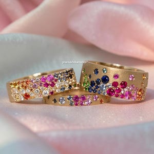 Pode incluir: Três anéis de ouro com pedras preciosas coloridas. Um anel é coberto por uma variedade de pedras preciosas, enquanto os outros dois têm pedras espalhadas. Os anéis são colocados sobre um fundo rosa e branco.