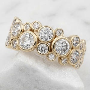 May include: A gold ring featuring a cluster of round, clear, faceted stones of varying sizes set in individual bezels. The band displays a textured, organic arrangement of stones across its surface, with the text Pramukhdiamonds visible.
