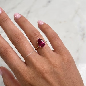 Op de afbeelding: Een gouden ring met een grote, peervormige, dieprode edelsteen. De ring is gezet op een slanke gouden band en wordt getoond op een hand met verzorgde nagels. Het ontwerp van de ring is eenvoudig en benadrukt de levendige kleur van de edelsteen.
