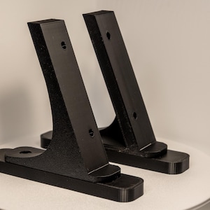 Könnte beinhalten: Zwei schwarze, abgewinkelte Halterungen mit rechteckiger Basis und gebogenem Träger. Jede Halterung hat zwei Löcher. Die Halterungen bestehen aus einem strukturierten Material und sind zur Unterstützung eines Objekts bestimmt.