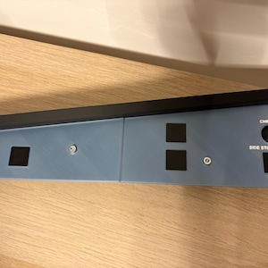 Pode incluir: Um painel de controlo retangular azul-claro com uma borda superior preta, vários botões quadrados pretos e um botão circular com a etiqueta CHRONO. O painel inclui o texto SIDE STICK PRIORITY impresso a branco perto de um botão preto e parafusos prateados.
