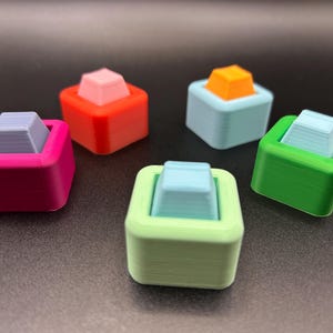 May include: Five small, square-shaped objects in various colours, each with a smaller square on top. The colours include pink, red, blue, green, and light green. The top squares are in shades of grey, orange, and pink.
