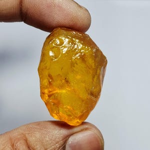 Peut inclure: Une pierre précieuse brute et translucide de couleur ambre avec une forme irrégulière à multiples facettes. La pierre présente une teinte chaude orange doré et une texture de surface polie et brillante qui capte la lumière, révélant des variations internes.