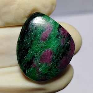 Könnte beinhalten: Ein polierter, tropfenförmiger Edelstein mit einer auffälligen Farbmischung. Der Stein zeigt eine satte grüne Basis mit markanten Flecken von tiefroten und schwarzen Einschlüssen. Die Oberfläche ist glatt und reflektierend, wodurch die natürlichen Muster hervorgehoben werden.