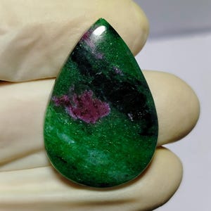 Może przedstawiać: Wypolerowany kamień szlachetny w kształcie łzy, o żywej zielonej podstawie z uderzającymi wtrąceniami głębokiej czerwieni i czerni. Powierzchnia kamienia jest gładka i odbijająca światło, ukazując naturalne wzory i kolory.