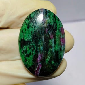Peut inclure: Pierre précieuse ovale avec un mélange de vert, de noir et de magenta. La surface polie reflète la lumière, révélant les motifs et textures naturels. La pierre est tenue par une main gantée.
