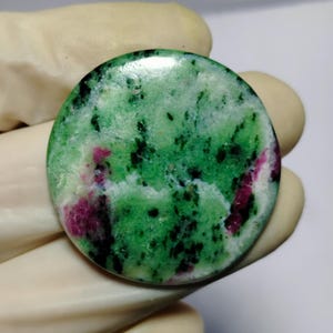 Może przedstawiać: Okrągły, wypolerowany kamień szlachetny o żywym zielonym kolorze podstawowym, nakrapiany czernią i plamami głębokiej czerwieni. Kamień ma gładką, błyszczącą powierzchnię i jest trzymany przez dłoń w rękawicy. Kamień szlachetny to minerał naturalny.