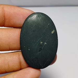 May include: An oval-shaped, dark green gemstone with light-colored veining and small flecks. The polished surface reflects light, highlighting the natural patterns within the stone. The gemstone is held in a hand.
