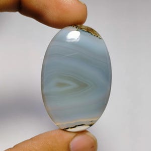 May include: An oval-shaped, polished gemstone with a light blue and white banded pattern. The stone has a central eye-like design and a brown and black edge. The gemstone is held between two fingers.