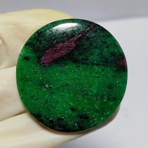 Peut inclure: Une pierre précieuse ronde et polie, avec une base verte vibrante, parsemée de noir et une proéminente veine magenta foncé. La surface de la pierre est lisse et réfléchissante, révélant des motifs naturels.